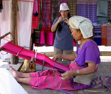 A village lady in Thailand, weaves cloth while a tourist takes a photo.